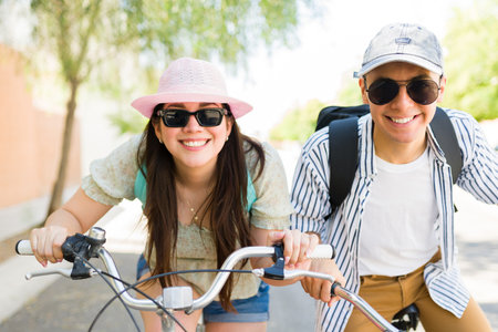 Fun girlfriend and boyfriend with sunglasses and hats smiling making eye contact and laughing during a bike ride outdoors during summerの写真素材