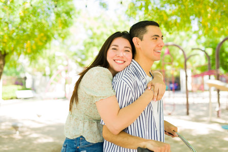 Beautiful caucasian woman hugging her happy boyfriend and looking at the camera while riding a bike together in the parkの写真素材