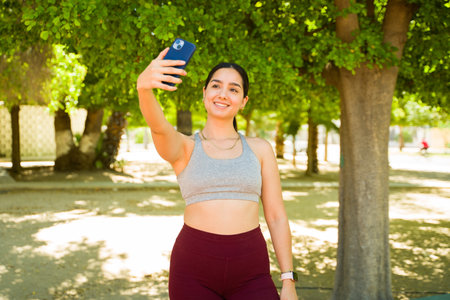 Beautiful latin woman in activewear smiling taking a selfie with her smartphone to post on social...