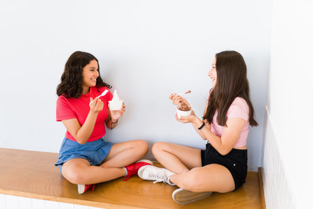 Hispanic teenager girls talking about gossip and smiling looking happy while eating delicious ice cream or frozen yogurtの写真素材