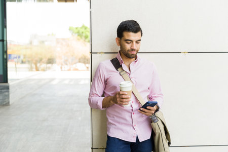 Business man texting on his smartphone carrying a messenger bag and drinking coffee while walking on his commute to workの写真素材