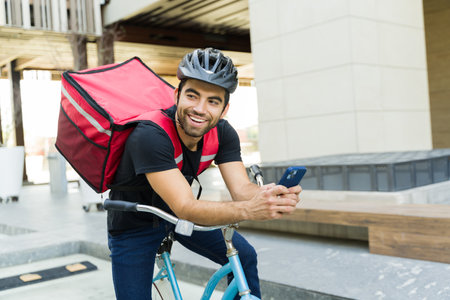 Cheerful latin worker smiling while riding his bicycle and working in the food delivery service carrying a backpackの写真素材