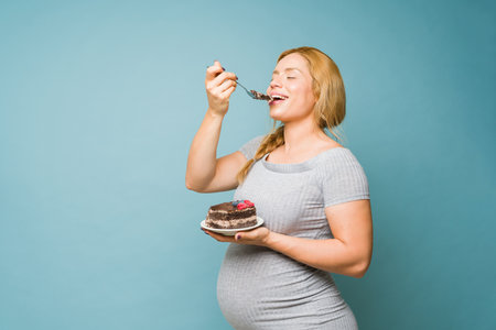 Attractive pregnant woman eating a big slice of cake with her eyes closed in a studioの写真素材