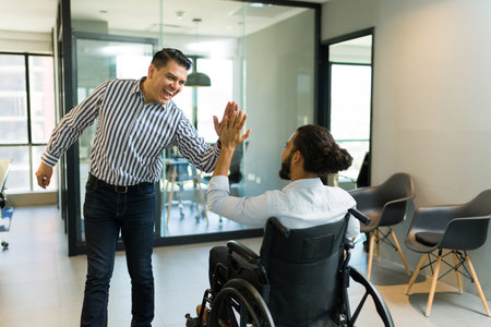 Cheerful mid adult businessman giving high five to a disabled colleague on wheelchair in officeの写真素材