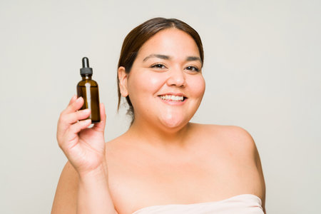 Cheerful fat latin woman smiling while showing a bottle of hyaluronic acid product while using beauty skin care productsの写真素材