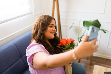 Attractive hispanic woman smiling receiving flowers from her long-distance boyfriend and taking a selfie with her smartphone for social mediaの写真素材