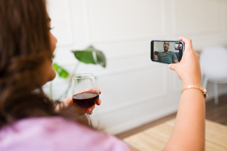 Hispanic woman drinking wine on a long distance relationship having a romantic virtual date with her boyfriend on a video callの写真素材
