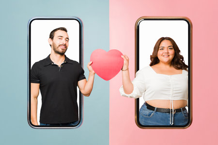 Smiling boyfriend and girlfriend having a love match on an online dating app and holding a heart through the smartphone screenの写真素材
