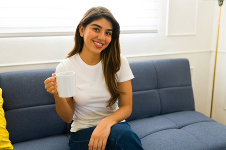 Beautiful happy woman drinking on a mock-up coffee mug and smiling wearing a white casual t-shirt at...
