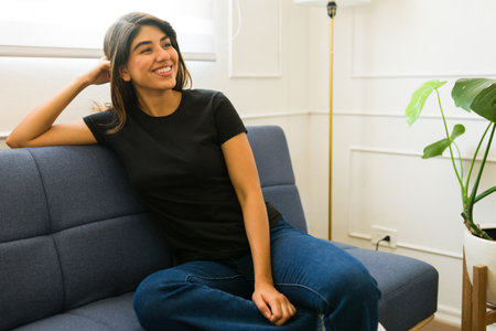 Cheerful relaxed woman sitting on the sofa at home wearing a black mockup t-shirt for a designの写真素材