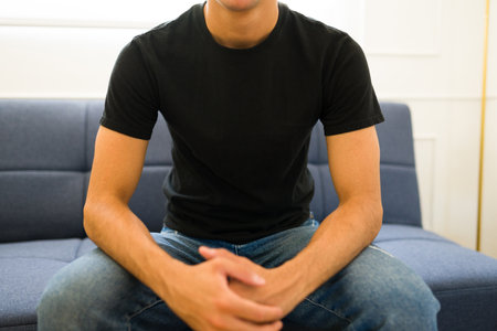 Young man sitting on the sofa wearing a black mock-up t-shirt ready for a copy space design productの写真素材