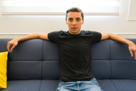 Portrait of a handsome latin young man relaxing on the sofa while wearing a casual black mock-up...