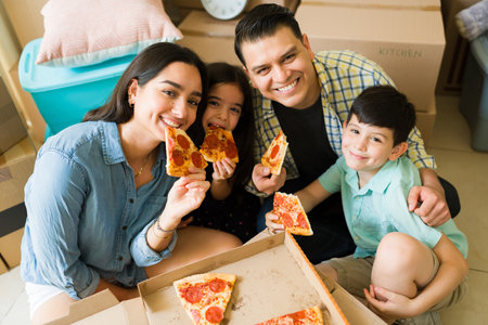 Happy family of four smiling looking at the camera while eating pizza for dinner using cardboard boxesの写真素材