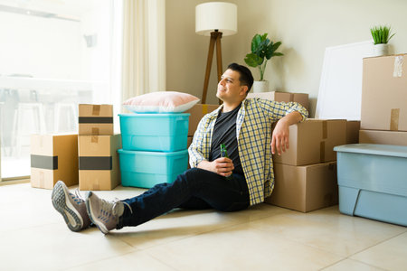 Relaxed independent man drinking a beer looking happy while unpacking boxes and moving to a new...