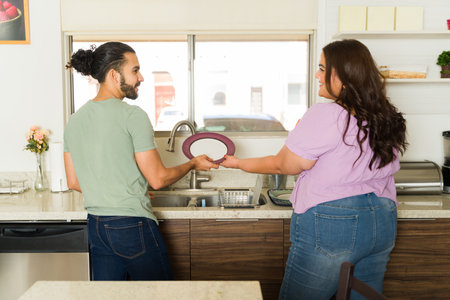 Rear view of a happy romantic couple smiling enjoying washing the dishes in the kitchen sink while doing house chores togetherの写真素材
