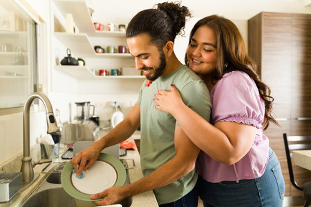 Romantic happy boyfriend and girlfriend smiling and hugging while enjoying doing house chores and domestic life while washing the dishes in the kitchenの写真素材