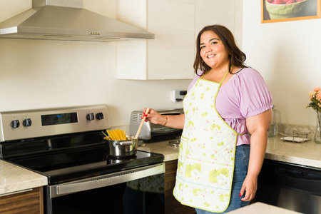 Beautiful happy fat woman wearing an apron while cooking pasta in the electric stove and enjoying...