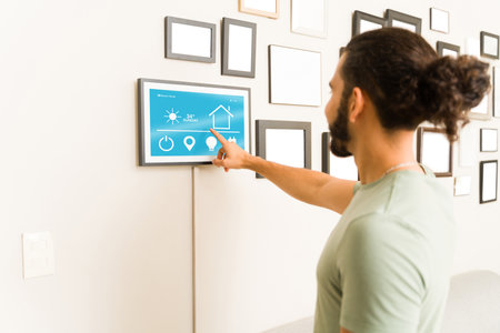 Mexican young man seen from behind using the tablet to control the air conditioning and lighting for...