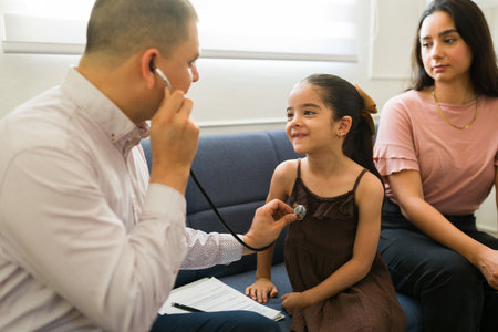 Latin pediatrician doing a patient examination with a stethoscope during a doctor's visit at home because of a sick little kid and her momの写真素材