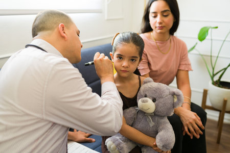 Beautiful sick little kid hugging her teddy bear while having a doctor's visit using an otoscope because of an ear infectionの写真素材