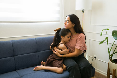 Beautiful worried mother hugging her sick little daughter looking sad while sitting together on the couch at homeの写真素材