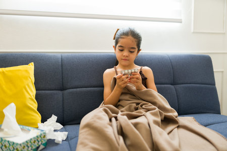 Adorable little girl with a blanket having health problems looking sick while taking medicine pills sitting on the couchの写真素材