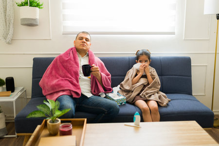 Father and daughter feeling ill and sick suffering from a bad cold or flu at home drinking hot tea and blowing their noseの写真素材