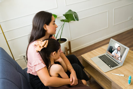 Hispanic mother hugging her sick little kid seen from behind while talking to a virtual doctor on the laptop during a video callの写真素材