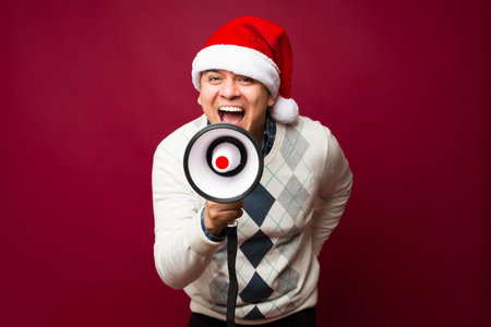 Mexican man in his 30s looking happy with a santa hat while screaming about the christmas holidays using a megaphoneの写真素材