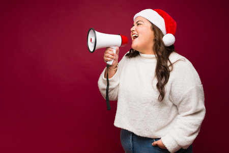 Profile of a beautiful obese woman with a santa hat screaming a christmas message using a megaphone looking excitedの写真素材