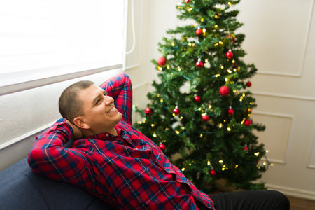 Relaxed happy latin man relaxing and smiling on the couch next to the christmas tree while enjoying the holidays at homeの写真素材