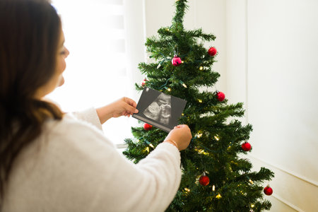 Latin plus size woman seen from behind putting a baby ultrasound on the christmas tree while happy about her pregnancy in the holidaysの写真素材