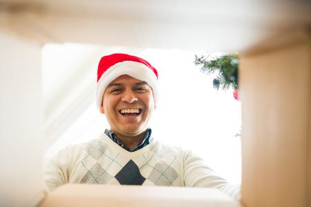 Excited latin man opening a christmas present and looking at the box from a low angle while celebrating christmas with a santa hatの写真素材