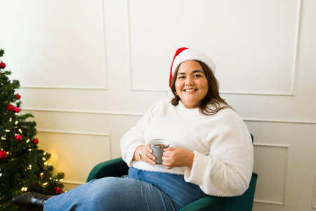 Happy fat hispanic woman smiling making eye contact with a santa hat drinking hot chocolate at home next to the christmas treeの写真素材
