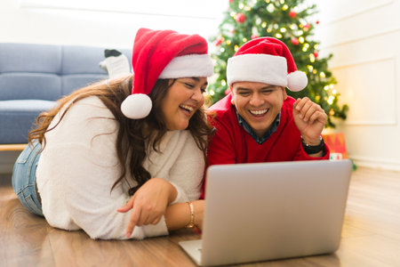 Excited latin couple laughing and having fun while online shopping on the laptop buying christmas presents for the familyの写真素材