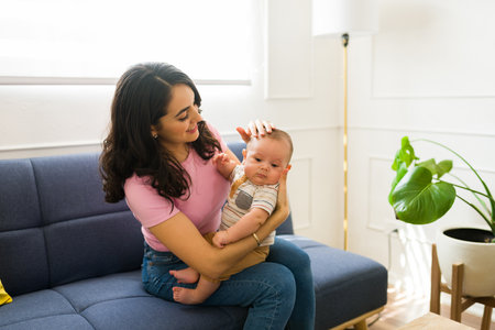 Beautiful happy mom at home smiling to her adorable baby while relaxing sitting on the couch in the...
