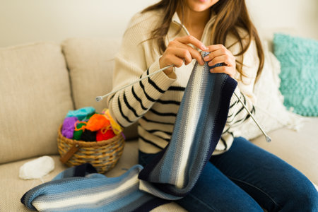Cheerful latin young woman enjoying her hobby and knitting a cozy scarf while relaxing on her cozy living room sofaの写真素材