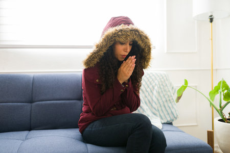 Cold young woman wearing a jacket and winter clothes blowing into her hands trying to warm up sitting on the couch without a radiatorの写真素材