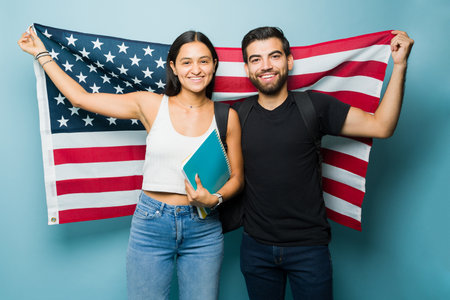 Hispanic students in their 20s holding a US flag while learning English and studying college against a blue studio backgroundの写真素材