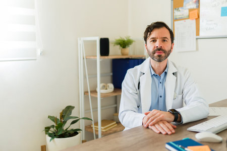 Attractive latin doctor looking at the camera wearing a lab coat sitting at his office desk waiting for a patientの写真素材
