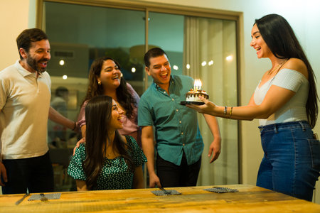 Excited group of friends singing happy birthday and bringing a surprise cake while celebrating...