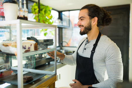 Attractive hispanic young man smiling working as a barista handing sweet dessert to a customer at...