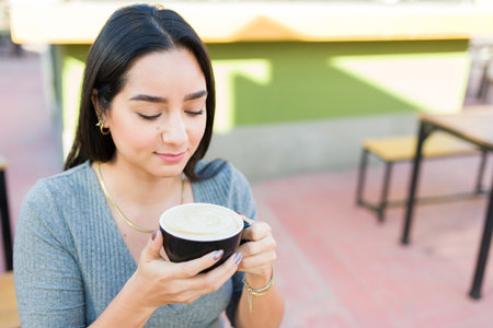 Beautiful hispanic young woman smiling while drinking cappuccino and sitting at the outdoor coffee shopの写真素材