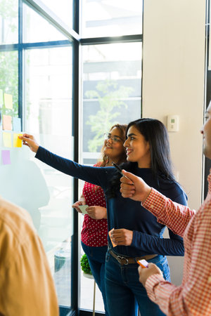 Diverse group of businesspeople using sticky notes on a glass wall to work on some team building...