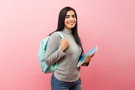 Hispanic university student smiling looking happy while studying college carrying her books and backpack in a pink backgroundの写真素材