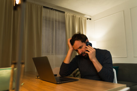 Stressed hispanic man looking worried while talking on the phone having problems and working at...