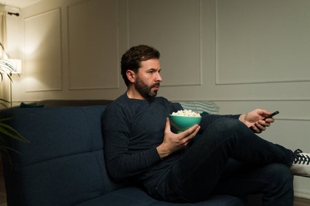 Handsome hispanic man relaxing on the couch while watching television or movies in the living room at night and eating popcornの写真素材