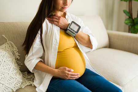 Caucasian expecting mother checking her high blood pressure with a monitor while showing her...