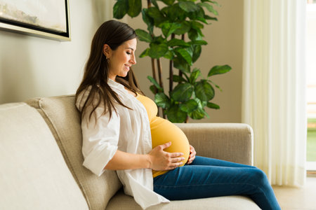 Beautiful expecting mother smiling while looking at her pregnant belly and relaxing in the living...