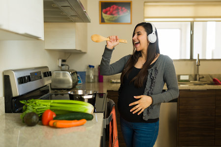 Excited pregnant woman listening to music with headphones while cooking a healthy lunch in the kitchen and enjoying her pregnancy having funの写真素材